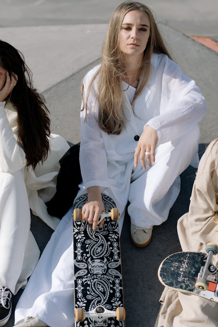 Woman In White Long Sleeve Clothes Sitting With Her Friends On Concrete Ground Holding Skateboards