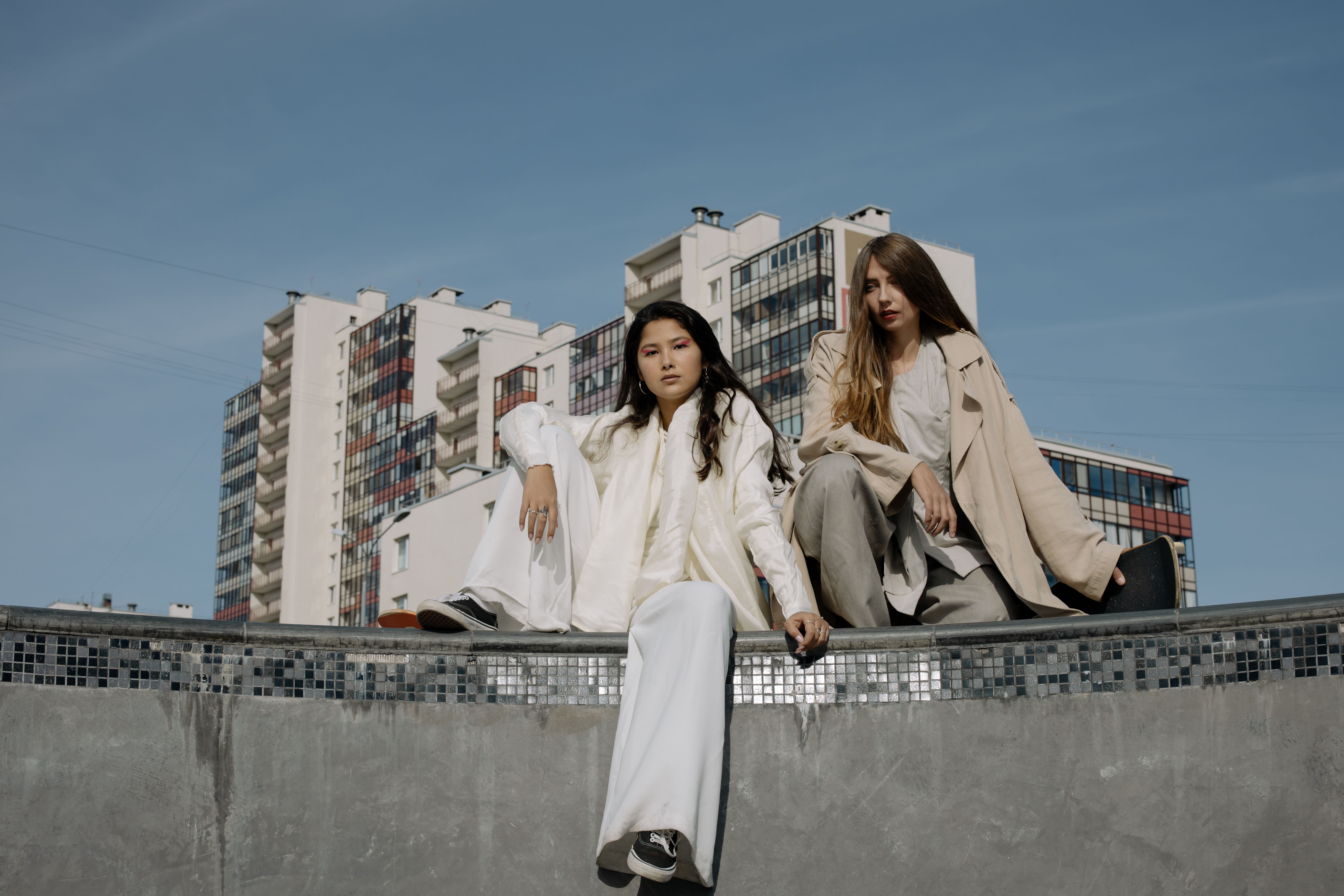 Two fashionable women sitting outdoors against a backdrop of urban buildings.