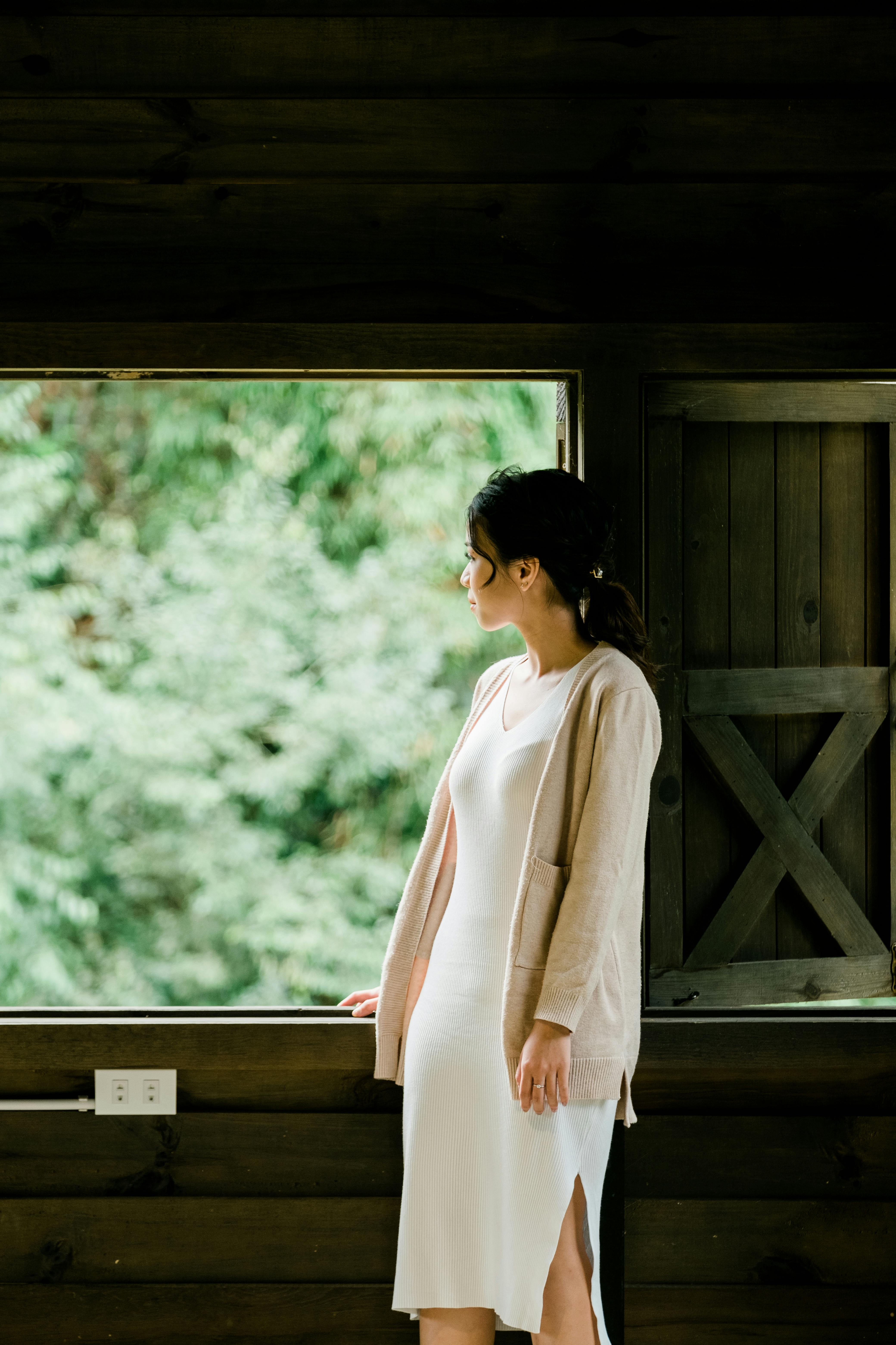 A woman gazes outside from a wooden house, dressed in a white dress and cardigan.