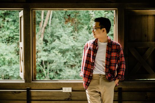 A man wearing glasses stands by an open window, gazing at the lush forest outside.