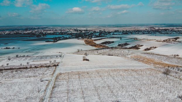 A stunning aerial view of snowy fields in Săcueni, Romania with a distant village under a blue sky.