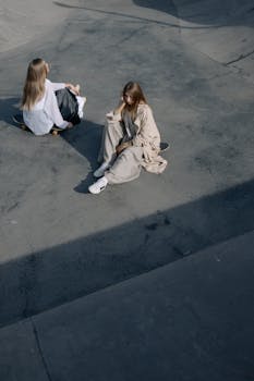 Two fashionable women sitting on skateboards in a skate park, exuding a relaxed and stylish vibe.