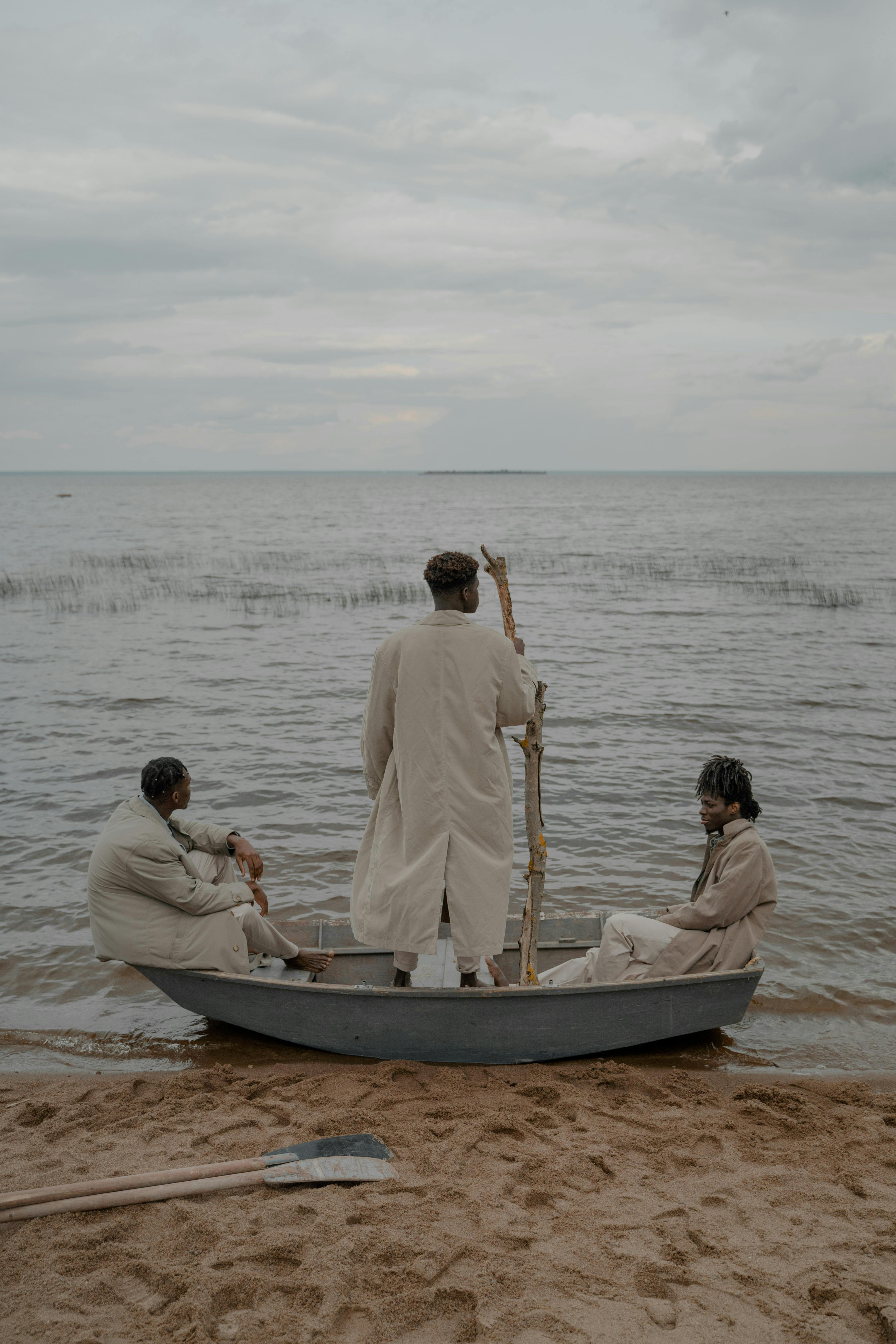 Three men wearing trench coats sitting and standing in a boat on a sandy beach with overcast skies.