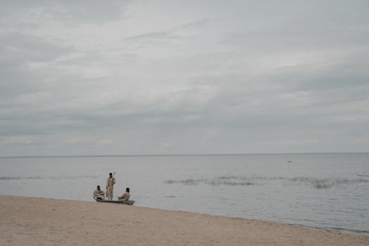 Three individuals in a wooden boat on a serene beach with overcast skies. Perfect for scenic coastal themes.