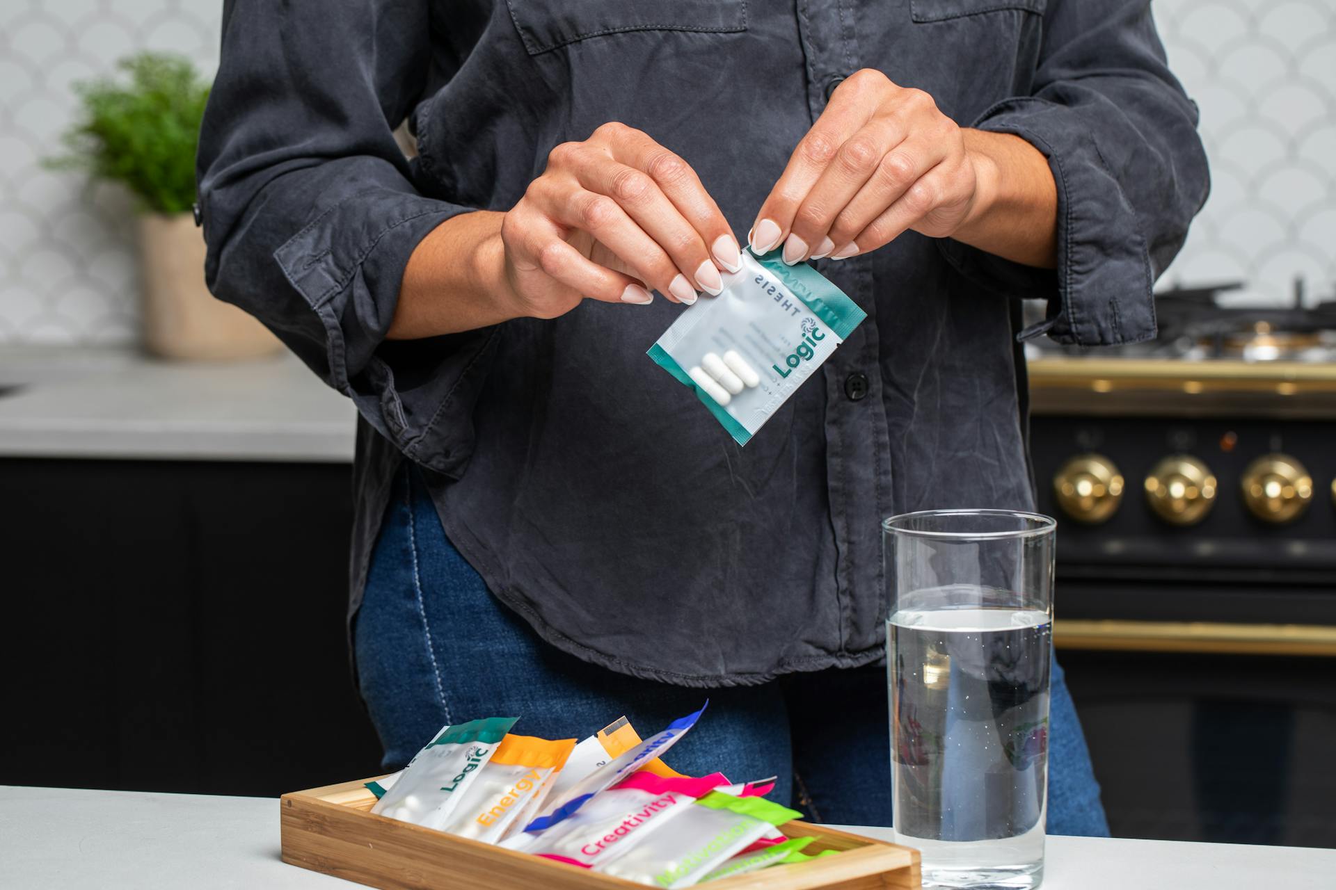 Close-up of hands holding a supplement package with pills next to a full glass of water in a kitchen setting.