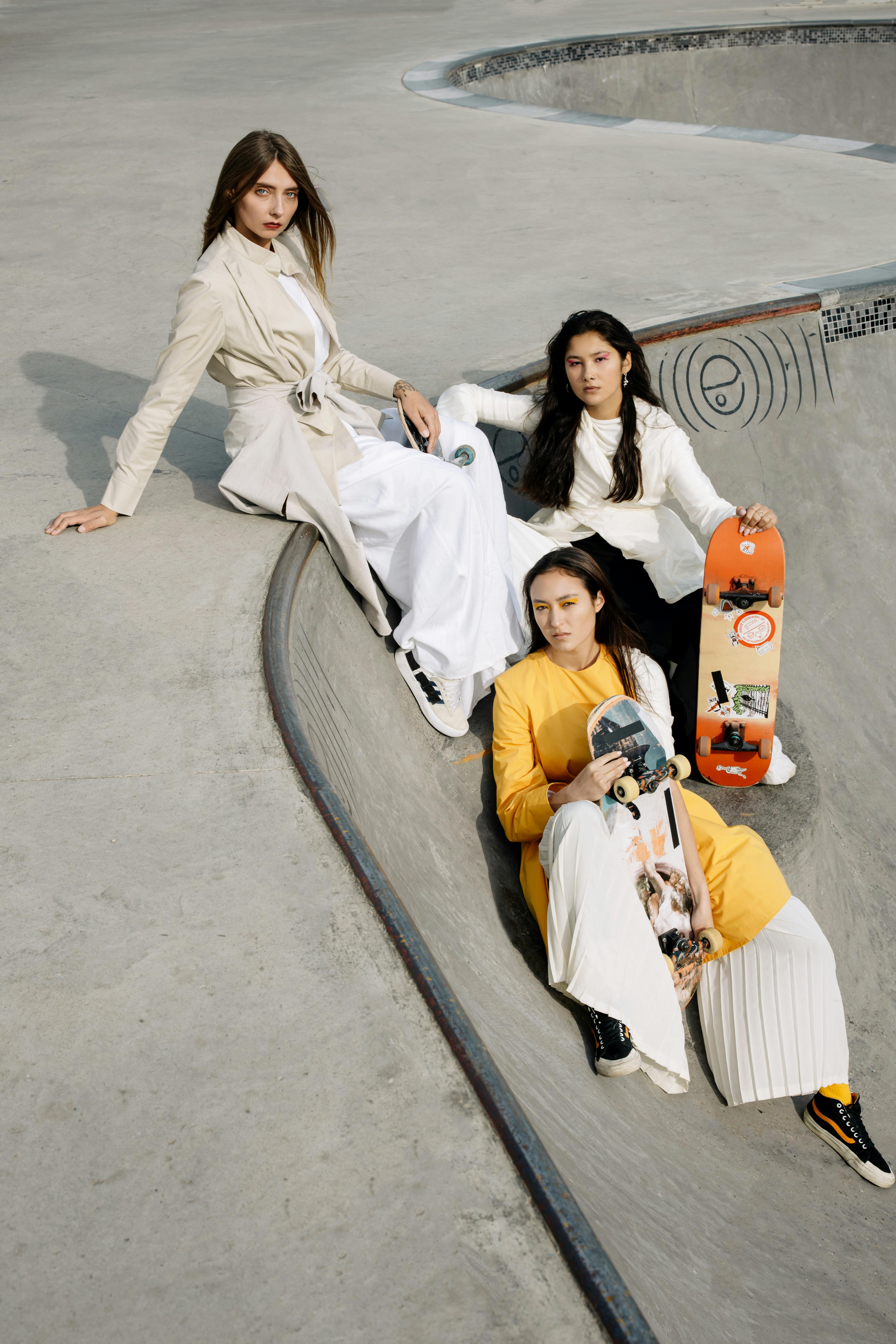 Three fashionable women pose with skateboards in an urban skate park setting.