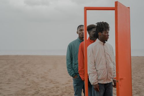 Free Three men stand in a line behind a red door on a beach, creating a surreal scene. Stock Photo