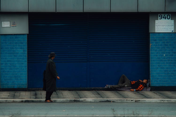 Woman In Black Clothes Walking On A Street And Man Sleeping Under Blue Garage Door