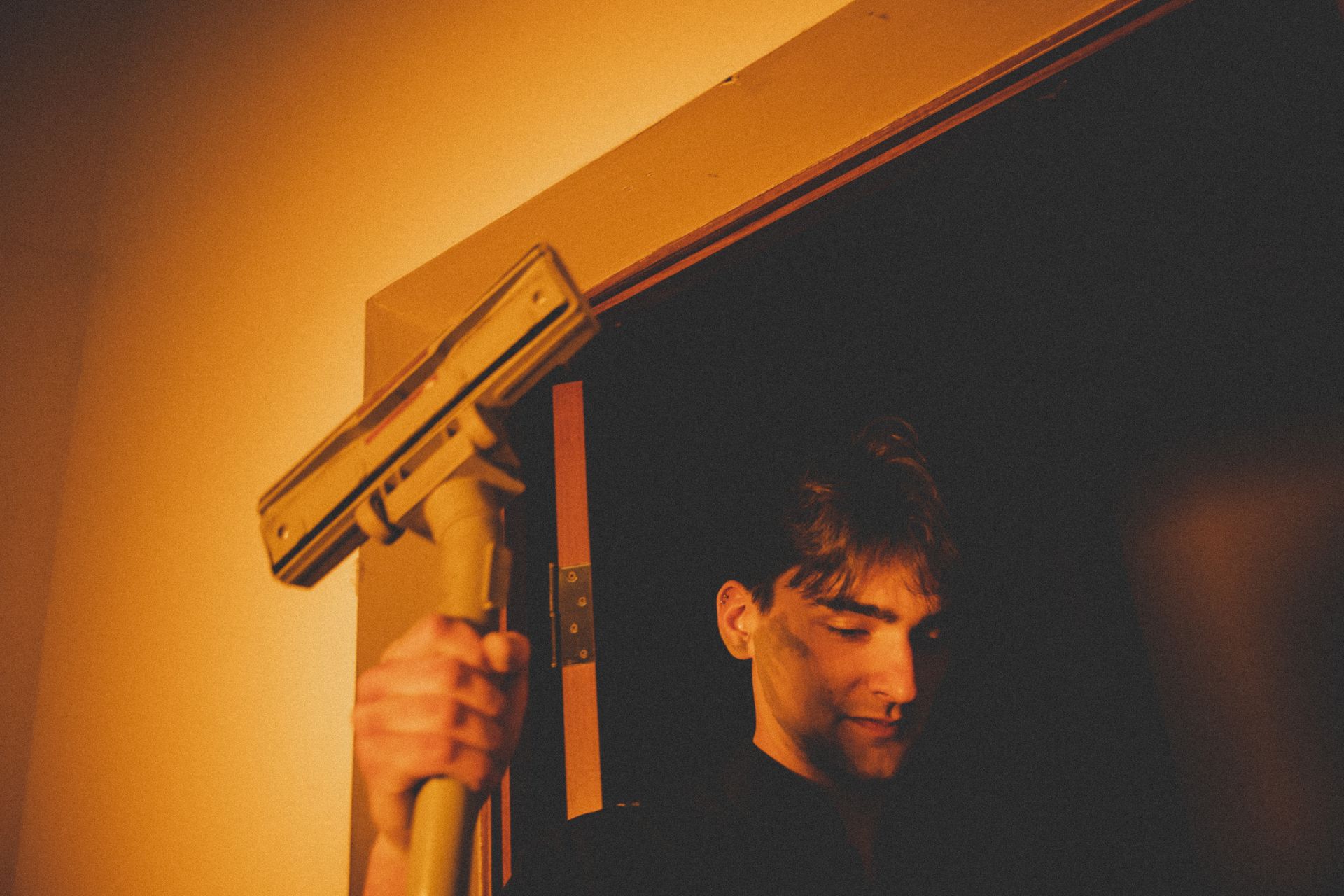 Young man standing in a dimly lit doorway, holding a vacuum cleaner, creating a moody atmosphere.
