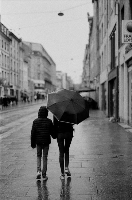 A mother and child walk under an umbrella on a rainy city street in black and white.