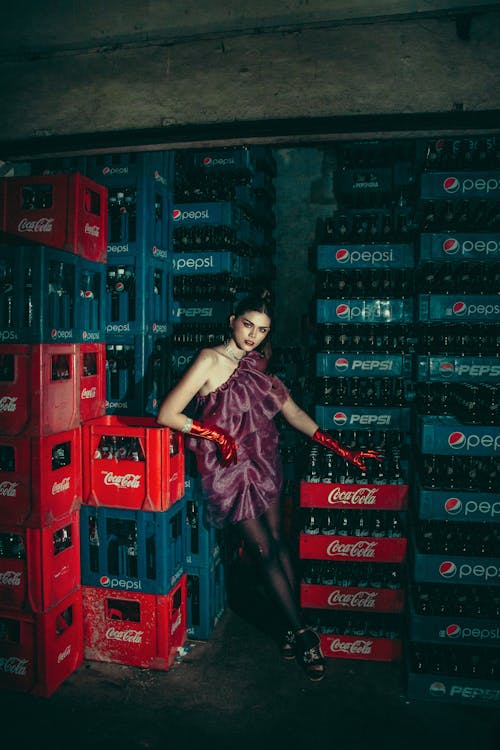 Woman in Purple Dress and Red Gloves Standing Among Crates of Soda