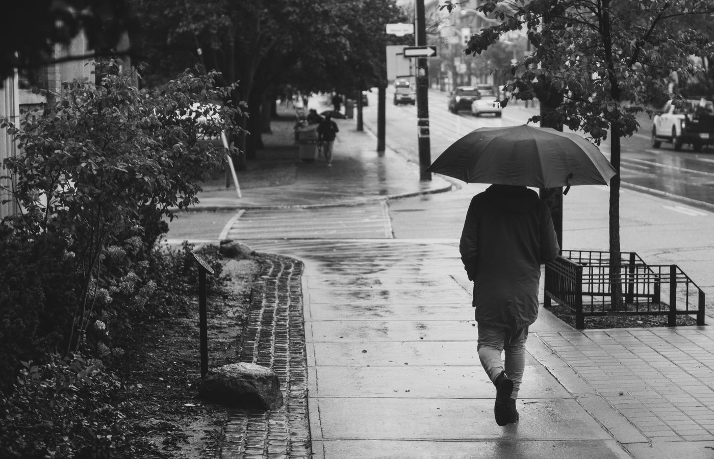A person walks on a rainy sidewalk holding an umbrella in a monochrome street scene.