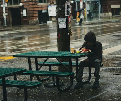 A solitary figure in a hoodie eats at a picnic table on a rainy day in the city.