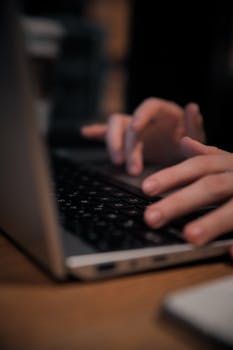 Blurred close-up shot of hands quickly typing on a laptop keyboard in a dimly lit setting.