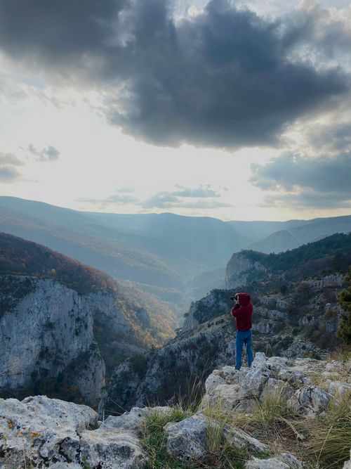 Free A photographer in a hoodie captures the breathtaking view of rocky mountains during sunset. Stock Photo