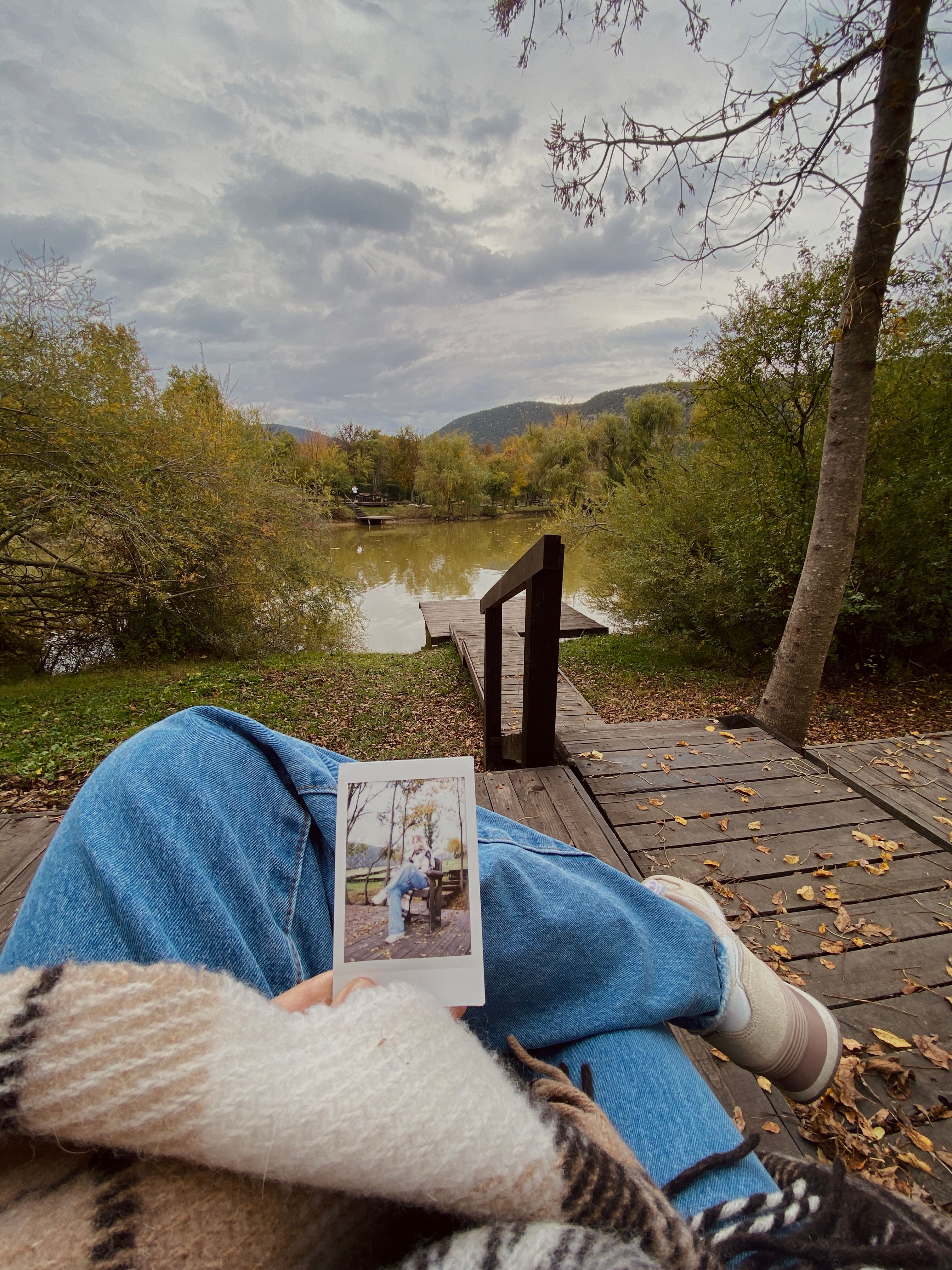 A serene lakeside scene with a person holding a polaroid picture, surrounded by autumn foliage.