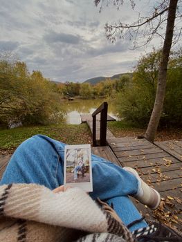 A serene lakeside scene with a person holding a polaroid picture, surrounded by autumn foliage.