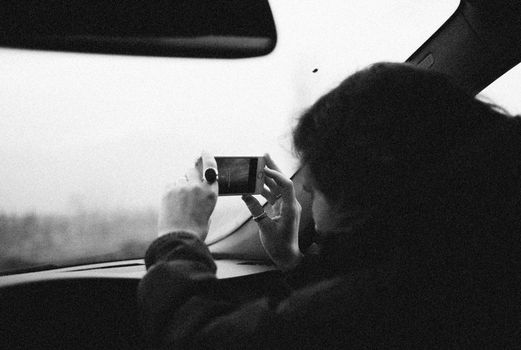Monochrome image of a person capturing a view with a smartphone from a car.