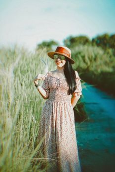 Woman in a floral dress, smiling with sunglasses and a hat, walking through nature on a sunny day.
