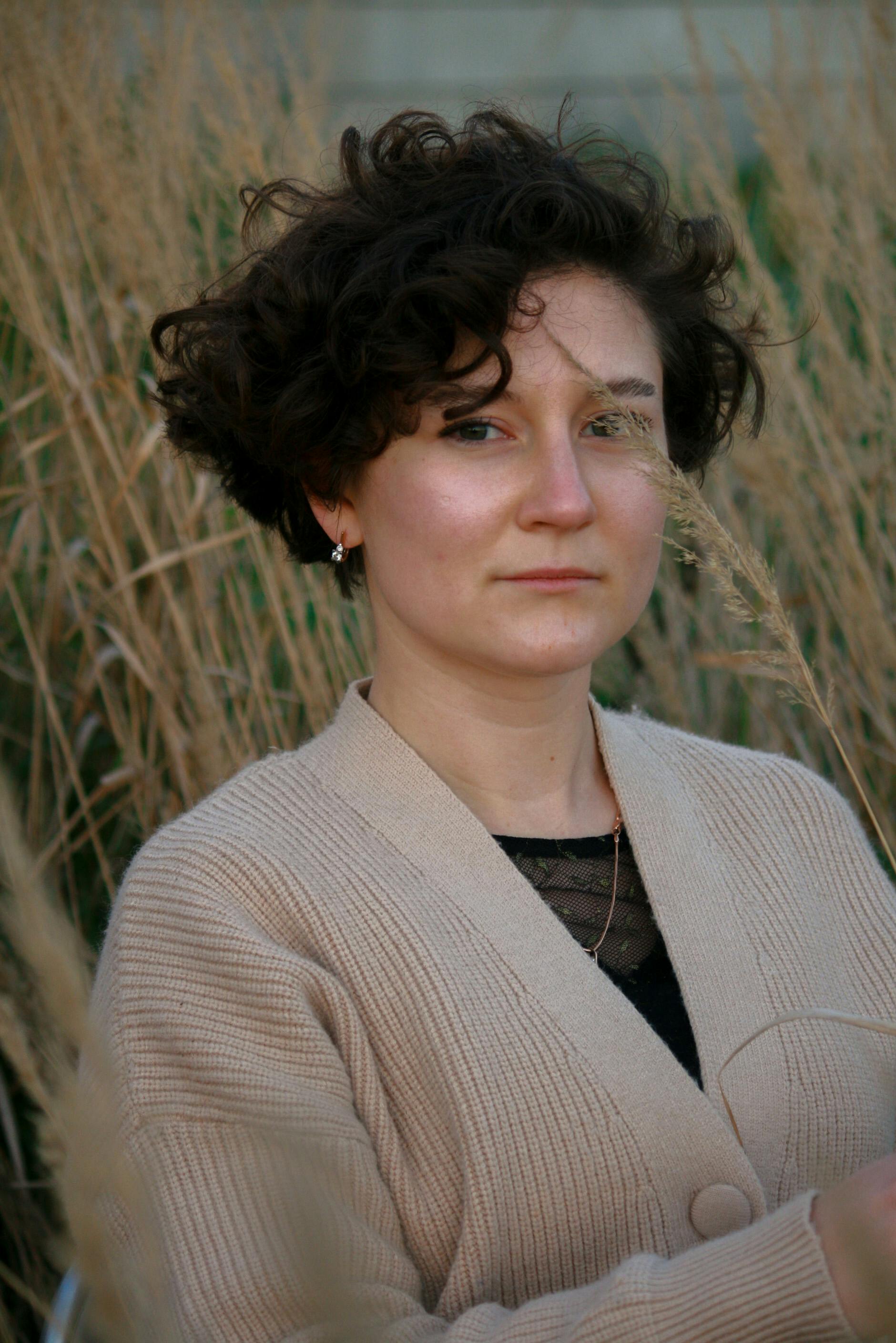 Portrait of a woman in a brown sweater standing in a grassy field, exuding a calm and natural ambiance.