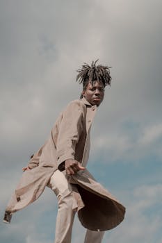 A fashionable man with dreadlocks wearing a trench coat, posing against a cloudy sky outdoors.