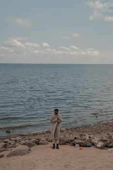 Man standing alone by a calm sea wrapped in a coat, suggesting a cool or cold atmosphere.