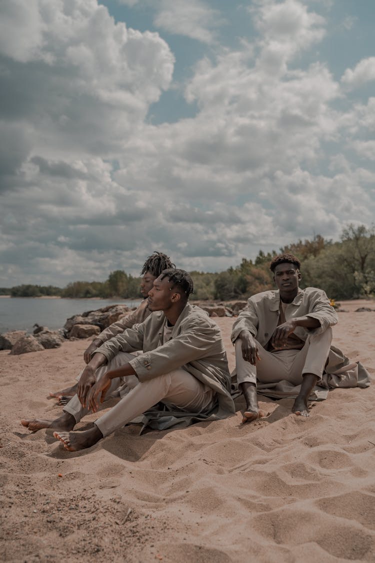 Small Group Of Dressed Up Men Sitting Barefoot On Sandy Beach On Sunny Day