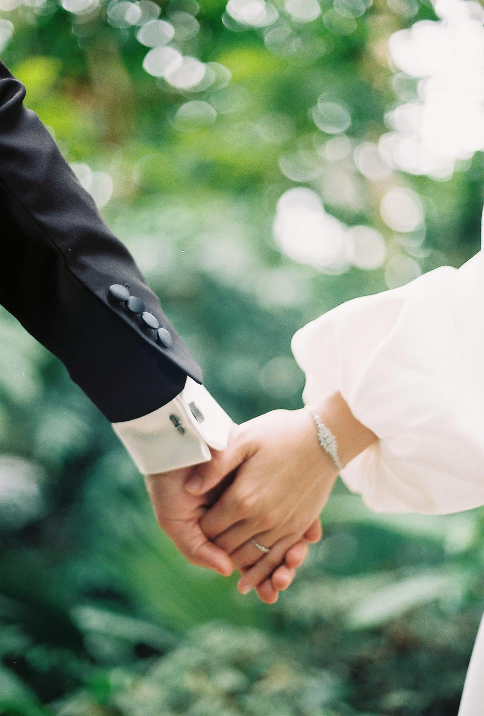 Close-up of a couple holding hands, dressed in elegant wedding attire, set against a lush green background.