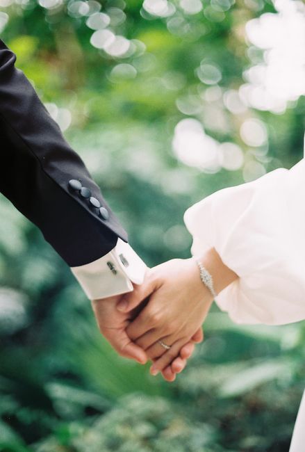 Close-up of a couple holding hands, dressed in elegant wedding attire, set against a lush green background.