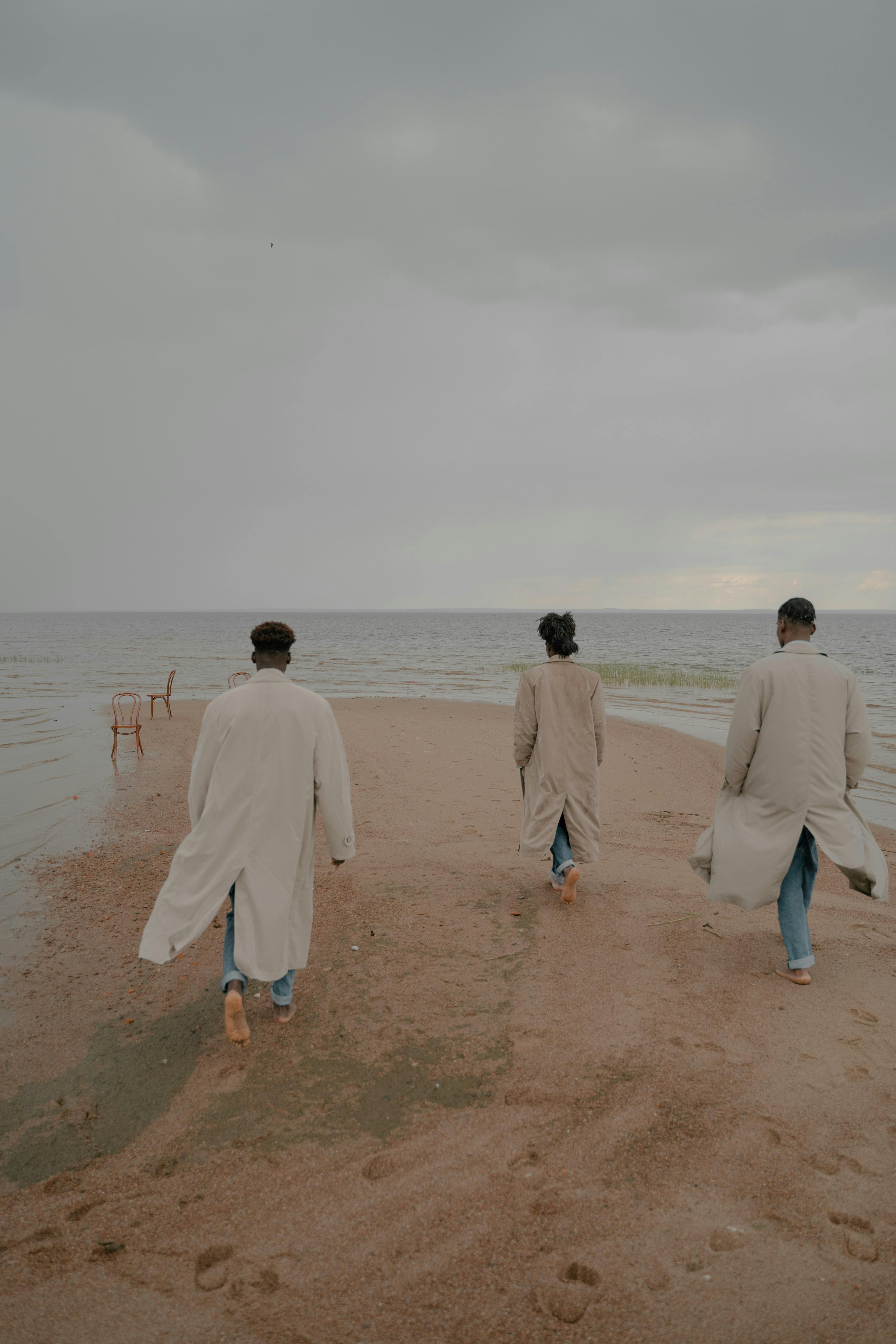 Three men in coats walking barefoot on a serene beach, evoking a tranquil autumn vibe.