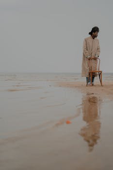 Conceptual photo of a man in coat standing beside a chair on a serene beach with reflection.