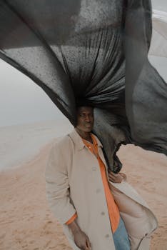 A fashionable man stands against a dramatic windy backdrop on a desolate beach.