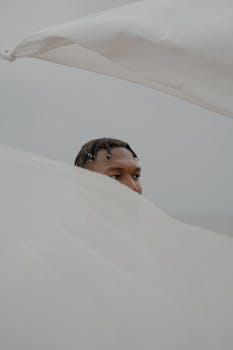 A partially obscured man with braids looks through soft, flowing fabric in a serene composition.