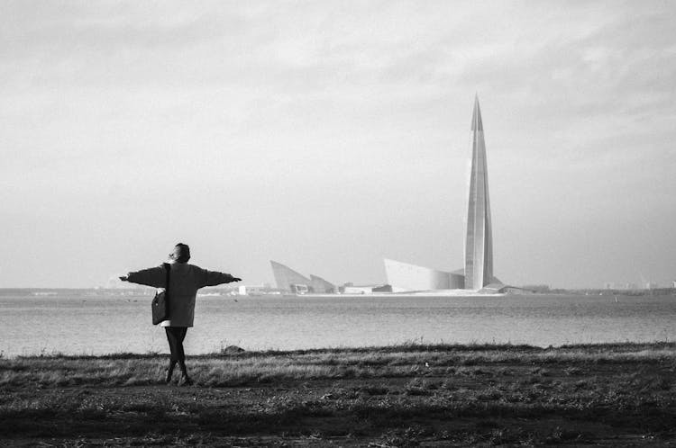 Black And White Photography Of A Woman With Stretched Hands On A Field And Modernist Church By The Sea