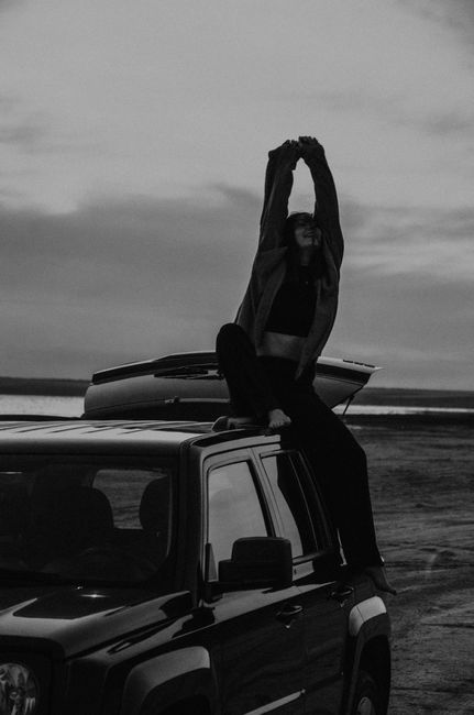 Monochrome image of a woman sitting on a car roof stretching her arms against a dramatic sky.