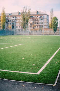 An empty soccer field with artificial turf and apartment buildings in the background, under a white sky.