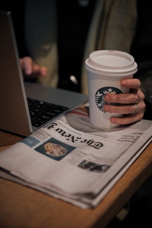 Free A woman holds a coffee cup while working on a laptop and reading the newspaper. Stock Photo
