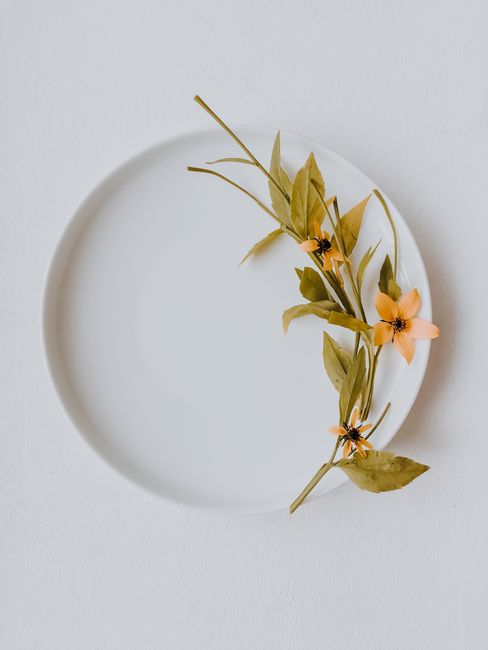 Minimalist floral arrangement with leaves and flowers on a white plate in a top view shot.