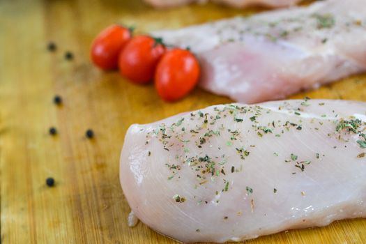 Fresh uncooked chicken breasts sprinkled with herbs, accompanied by cherry tomatoes on a wooden board.