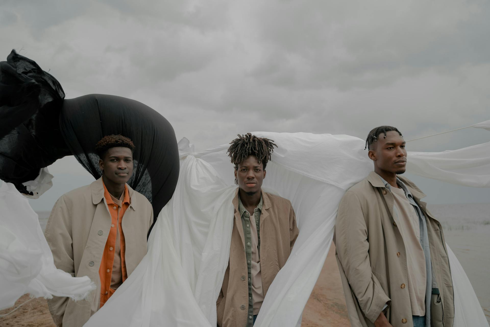 Men in Coats Standing on Beach With White and Black Cloth in Wind