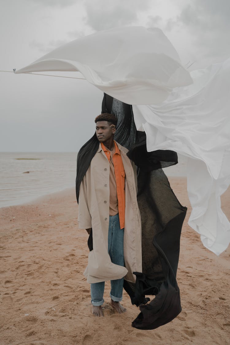 Man In Coat Standing On Beach With White And Black Cloth In Wind