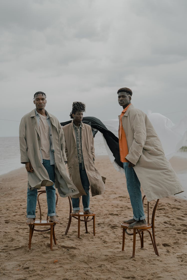 Men In Coats Standing On Chairs On Beach With White And Black Cloth In Wind