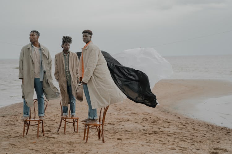 Men In Coats Standing On Chairs On Beach With White And Black Cloth In Wind