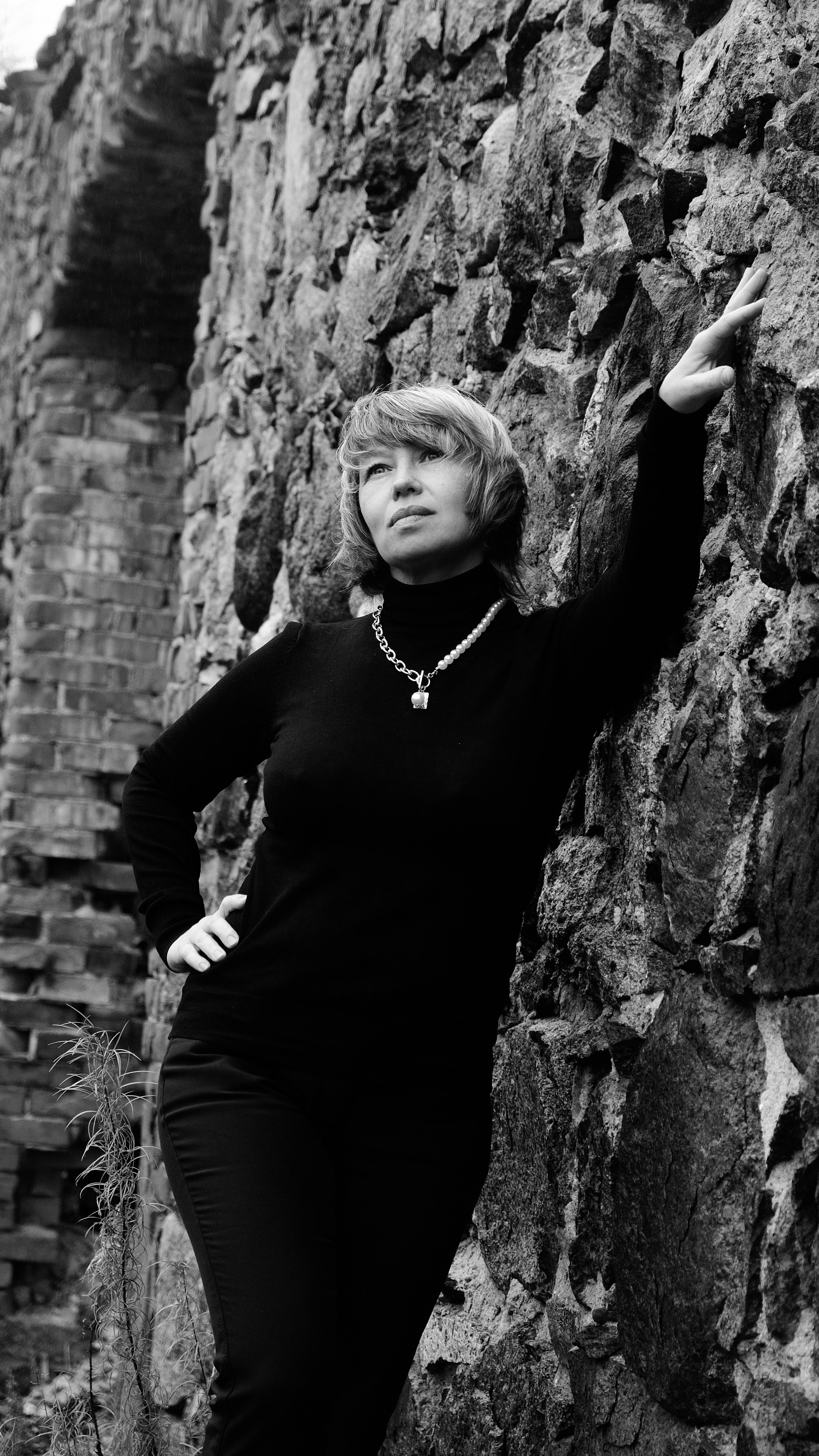 Grayscale portrait of a woman posing confidently against a rugged stone wall.