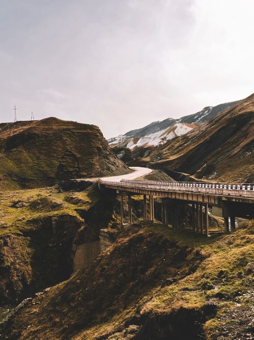 Free Scenic view of a bridge crossing rugged mountain landscapes with lush greenery and snow patches. Stock Photo