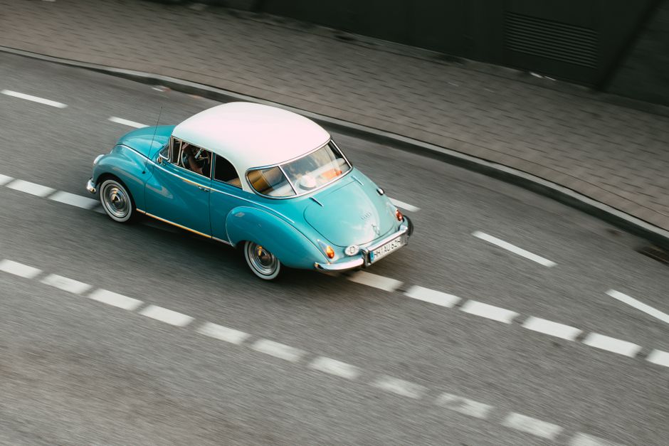 A teal classic car driving on a city road, captured in a high-angle shot for a vintage aesthetic.