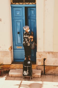 A woman wearing a face mask stands with luggage outside a blue door on a sunny street in Malta.