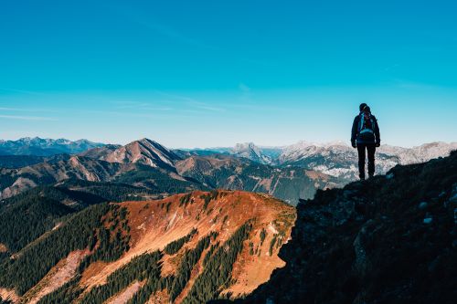 Free A lone hiker stands atop a mountain, taking in the breathtaking vista of mountain ranges under a clear blue sky. Stock Photo