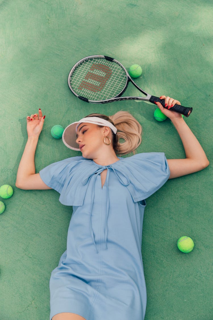 Woman In Blue Dress Laying On Ground Holding Racket Surrounded By Tennis Balls 