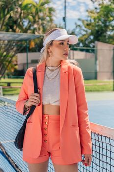 Stylish woman in coral blazer and shorts posing on tennis court, embodying sporty elegance.